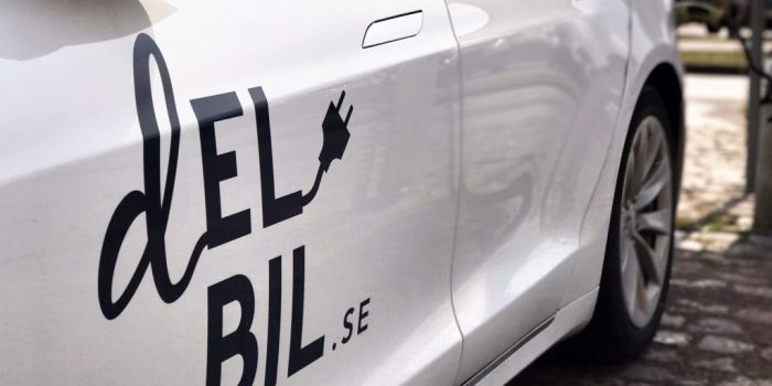 Närbild av en vit elbil med texten 'Delbil.se' och en laddkontakt som en del av logotypen. Bilen står parkerad på en stenlagd yta.