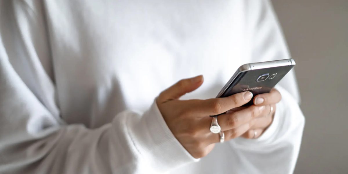 Person i vit långärmad tröja håller en smartphone med båda händerna. Fokus ligger på telefonen och händerna, medan bakgrunden är suddig.