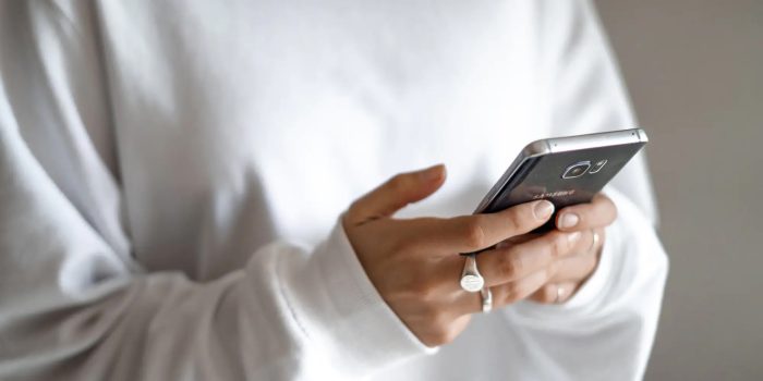 Person i vit långärmad tröja håller en smartphone med båda händerna. Fokus ligger på telefonen och händerna, medan bakgrunden är suddig.