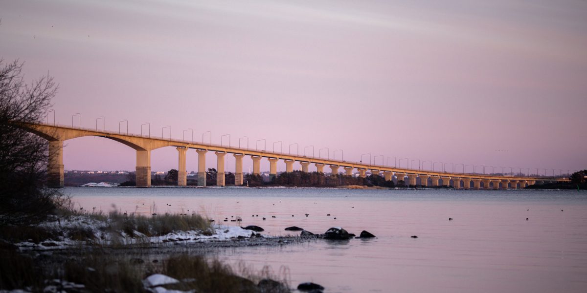 Ölandsbron i vinterljus, sedd från land med snötäckt strand i förgrunden och stilla vatten under bron.