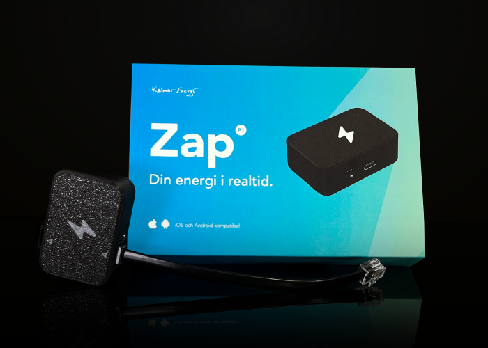 En svart realtidsmätare med blixtsymbol och kabel ligger framför en blågrön kartong med texten ”Zap – Din energi i realtid”. Kartongen visar en bild på produkten.