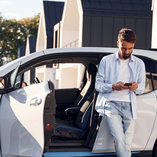 Person står lutad mot en vit elbil med öppna dörrar och tittar på en mobiltelefon. Bilen är parkerad på en uppfart med moderna hus i bakgrunden.