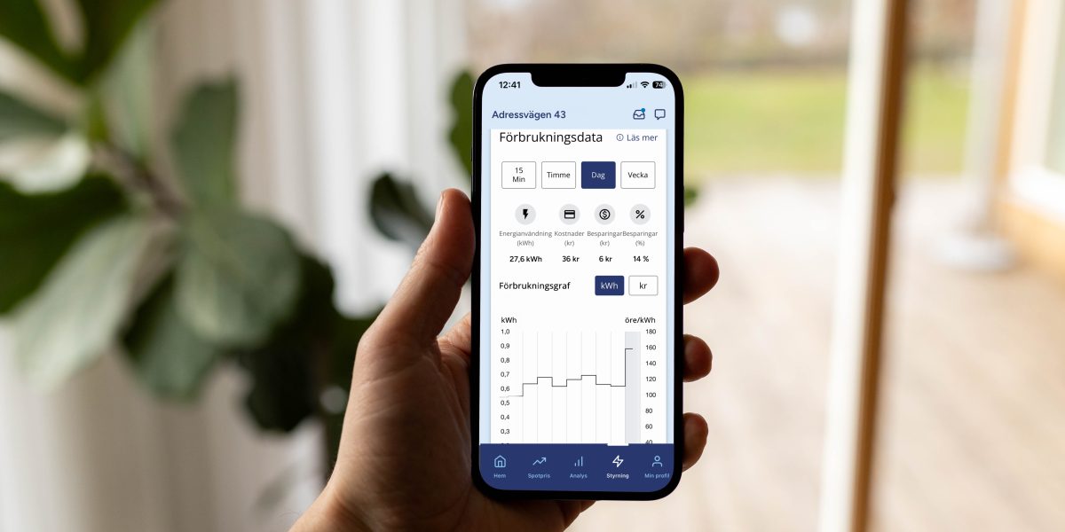 En hand håller en smartphone som visar Kalmar Energis app med förbrukningsdata för energianvändning. Skärmen visar en graf över förbrukning i öre per kWh och olika menyval för tidsintervall.