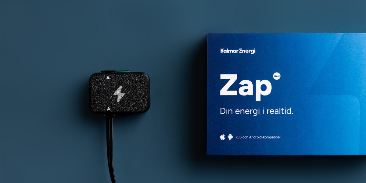 En svart realtidsmätare med blixtsymbol och kabel ligger framför en blå kartong med texten ”Zap – Din energi i realtid”.