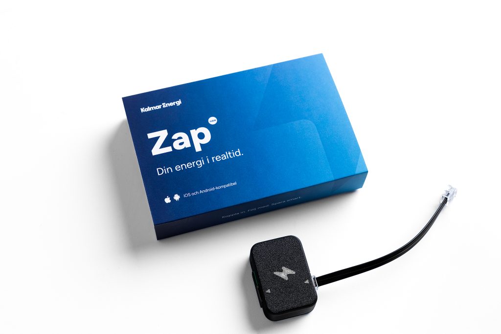En svart realtidsmätare med blixtsymbol och kabel ligger framför en blå kartong med texten ”Zap – Din energi i realtid”.