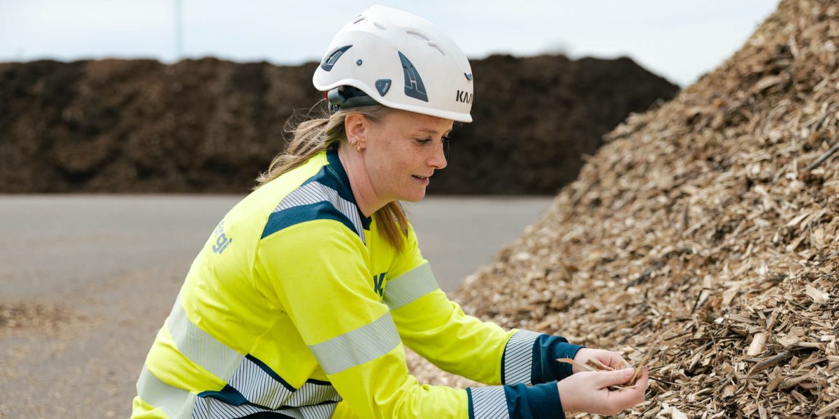 Medarbetare på Kalmar Energi i varselkläder och skyddshjälm sitter vid en stor hög med träflis och håller flisbitar i händerna utomhus på Moskogen.